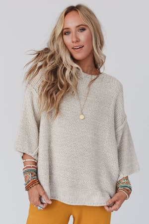 A woman with long, wavy blonde hair wears the Crisp Air Sweater in oatmeal, styled with mustard yellow pants, layered beaded bracelets, and a gold pendant necklace, standing against a plain light background.
