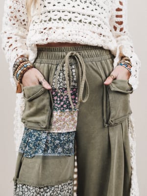 Person wearing a white lace top with green patchwork pants against a neutral background