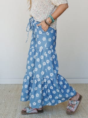A side-profile shot of a tiered maxi skirt in chambray blue with bold retro daisy print, styled with a multicolor speckled knit top, stacked boho bracelets, and beaded fringe sandals — the kind of outfit that feels equally at home at a farmers market