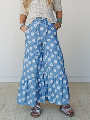 Tiered wide-leg pants in a chambray blue with an all-over retro daisy print, styled with a multicolor speckled knit top, beaded bracelets, and embellished sandals — the kind of outfit that looks like it was put together effortlessly but absolutely wa