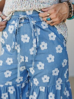 A close-up of chambray wide-leg pants covered in retro daisy appliqués, paired with a multicolor crochet top and stacked boho jewelry — the kind of outfit that feels like it was made for wandering somewhere sunny.