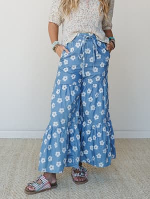 A breezy, retro-leaning wide-leg pant in chambray blue with bold daisy print, styled with a multicolor speckle knit top and embellished sandals — the kind of outfit that looks intentional without trying.