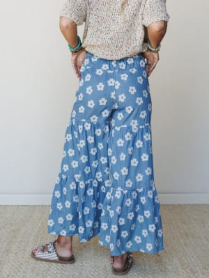 A flowy, wide-leg denim pant covered in retro daisy print is styled from behind with a multicolor knit top, beaded bracelets, and embellished sandals — casual and playful with a serious 70s energy.