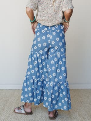 A flowy, wide-leg denim pant covered in retro daisy print is styled from behind with a multicolor knit top, beaded bracelets, and embellished sandals — casual and playful with a serious 70s energy.
