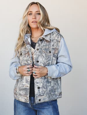 A relaxed, effortlessly cool hooded denim jacket in a washed leaf-print camo body with heathered grey fleece sleeves and hood — styled over a dark tee and jeans with layered silver and turquoise jewelry for that lived-in, expressive everyday look.