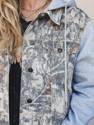 A close-up torso shot of a camo-leaf printed denim jacket with contrast light wash hoodie sleeves and drawstring detail, layered over a black tee with gold chain necklaces peeking out — the kind of jacket that looks like you found it somewhere cool b