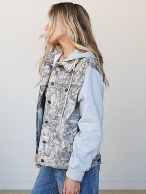 A side-profile shot of a mixed-media jacket that pairs a nature camo-print denim body with heathered gray fleece sleeves and a built-in drawstring hood — the kind of piece that looks like it got lost between a hiking trail and a weekend bonfire, in t