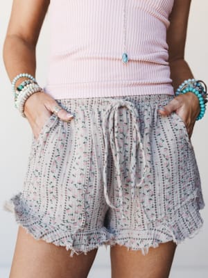 A close-up mid-shot of floral ruffle shorts in a soft taupe-and-pink ditsy print, styled with a ribbed pink tank and stacked turquoise and shell bracelets — the kind of effortless summer outfit you'd wear to the farmers market and never want to take