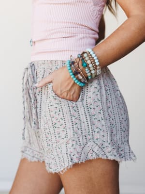 A close-up side shot of floral ruffle shorts paired with a pink ribbed top and a stacked mix of turquoise and gemstone bead bracelets — soft, summery, and the kind of outfit that feels as good as it looks.