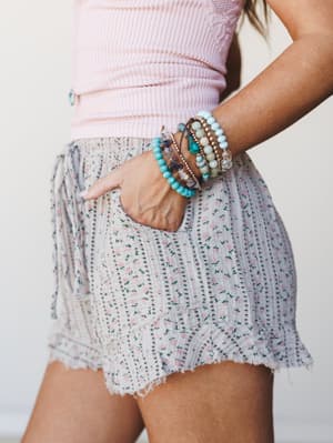 A close-up side shot of floral ruffle shorts paired with a pink ribbed top and a stacked mix of turquoise and gemstone bead bracelets — soft, summery, and the kind of outfit that feels as good as it looks.
