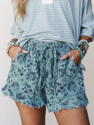 A close-up mid-shot of flowy floral gauze shorts in dusty turquoise paired with a striped top and stacked beaded bracelets — the kind of effortless warm-weather outfit you reach for without thinking twice.