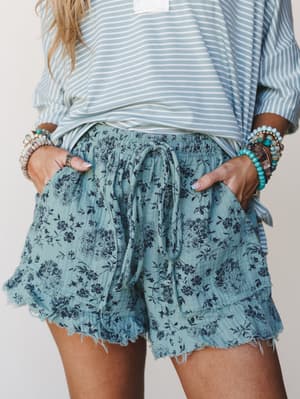 A close-up mid-shot of flowy floral gauze shorts in dusty turquoise paired with a striped top and stacked beaded bracelets — the kind of effortless warm-weather outfit you reach for without thinking twice.