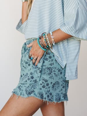 A close-up lifestyle shot of a floral ruffle-hem short in dusty turquoise paired with a striped bell-sleeve top, styled with a stack of beaded and stone bracelets and layered rings — easy, sun-warmed boho with a relaxed summer feel.