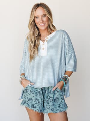 A breezy, easygoing outfit pairing an oversized blue-and-white striped henley top with floral ruffle shorts — the kind of look that feels like summer weekends with zero effort required.