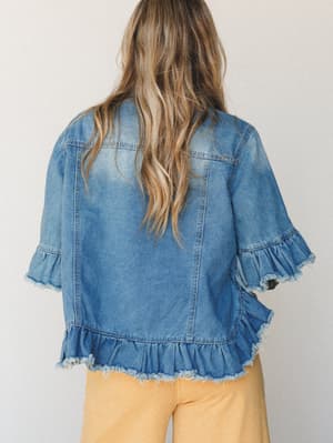 A back-facing shot of a ruffle-trim denim jacket in medium-light wash, styled with warm golden pants — the kind of piece that turns a regular Tuesday into something worth photographing.