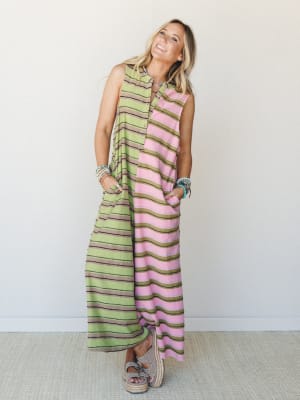 A flowy striped maxi jumpsuit in a playful mix of pink, green, and brown horizontal stripes, styled with stacked turquoise bracelets and platform espadrille sandals against a clean white wall — easy and expressive with a laid-back summer energy.