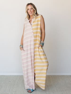 A relaxed, oversized sleeveless maxi jumpsuit in a soft colorblock stripe — blush pink on one side, warm mustard yellow on the other — styled with stacked turquoise bracelets and blue slide sandals against a clean white studio wall.