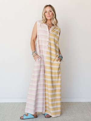 A breezy color-blocked maxi jumpsuit in soft pink and mustard stripes, styled with stacked beaded bracelets and sky blue sandals against a clean studio wall — the kind of outfit that feels like a slow morning somewhere warm.