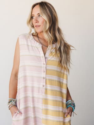 A relaxed sleeveless jumpsuit in a playful color-blocked stripe — blush pink on one side, mustard yellow on the other — styled with stacked turquoise and beaded bracelets and a delicate silver necklace for that effortless, well-traveled feel.
