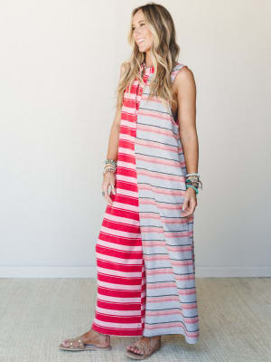 A striped maxi jumpsuit in a bold red-and-grey colorblock split — sleeveless, relaxed, and effortlessly summer — styled with stacked turquoise bracelets and flat sandals against a clean studio wall.