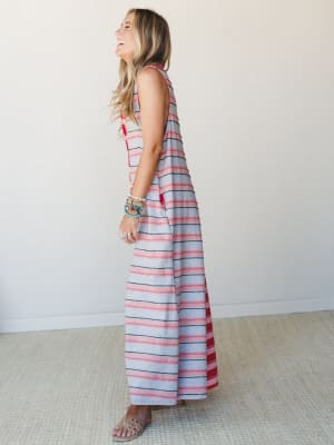 A laughing model shot in profile wears a flowy striped maxi jumpsuit in soft grey, blush pink, and red — styled with stacked boho bracelets and flat sandals, giving off that effortless summer energy.