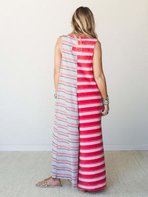 A back-facing model wears a bold color-blocked striped maxi jumpsuit in contrasting red and grey panels, styled with layered bracelets and studded sandals against a clean studio wall — effortless and expressive all at once.