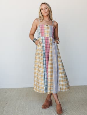 A breezy patchwork plaid midi dress in a mix of golden yellow, dusty rose, and lavender panels — worn with brown suede clogs and stacked bracelets, it feels like something you'd find at a vintage market and never want to take off.