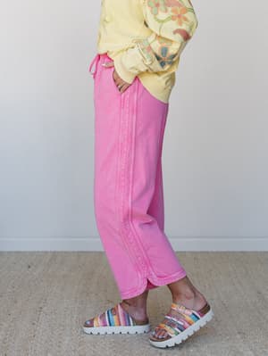 A side-profile shot of wide-leg cropped pants in a bold washed pink, styled with a floral embroidered yellow sweatshirt and multicolor crochet sandals — the kind of outfit that looks effortless but is clearly very intentional.