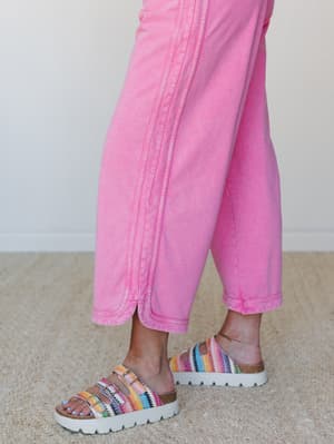 A close-up detail shot of wide-leg cropped pink denim pants with visible seam stitching, paired with multicolor crochet-strap platform sandals — playful and effortlessly lived-in.
