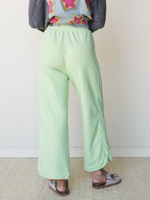 Shot from behind, a model wears wide-leg mint green sweatpants with a relaxed, lived-in feel — paired with a graphic appliqué jacket and patterned sandals for a playful, expressive layered look.