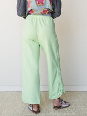 Shot from behind, a model wears wide-leg mint green sweatpants with a relaxed, lived-in feel — paired with a graphic appliqué jacket and patterned sandals for a playful, expressive layered look.