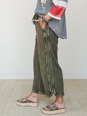 A side-profile shot highlighting acid-washed olive wide-leg crop pants styled with a striped color-block top, stacked beaded bracelets, and leopard print platform sandals — the kind of effortless, put-together look that feels like it came together on