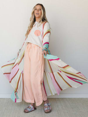 A bright, playful full-body shot of a model tossing open an oversized multicolor stripe mesh duster over a seashell graphic crop tee and wide-leg peach pants — the whole look reads like a beach vacation you've been daydreaming about since February.