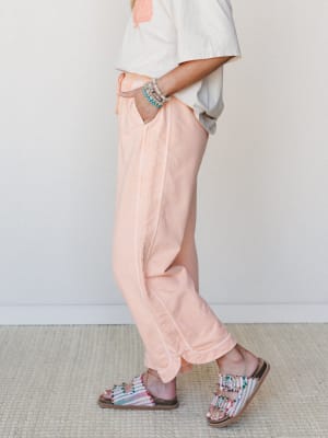 A side-angle mid-shot of relaxed, peach-toned wide leg pants styled with a cropped graphic tee, beaded bracelets, and colorful embellished sandals — easy, warm-weather dressing that feels intentional without trying too hard.
