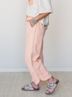 A side-angle mid-shot of relaxed, peach-toned wide leg pants styled with a cropped graphic tee, beaded bracelets, and colorful embellished sandals — easy, warm-weather dressing that feels intentional without trying too hard.