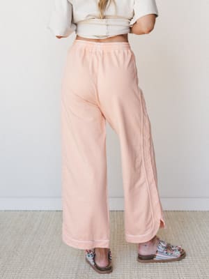 Soft peach wide-leg pants with contrast stitching and a banded hem are styled with a cream crop top and beaded sandals — the kind of easy, put-together look that works just as hard on a coffee run as it does on the couch.