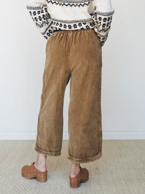 Shot from behind, a model wears wide-leg cropped pants in a warm washed brown paired with a cream fair isle sweater and tan studded clogs — an effortlessly cozy fall outfit with real lived-in energy.