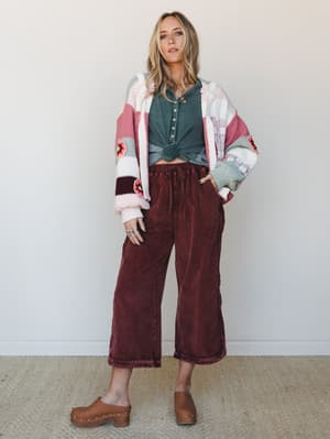 A relaxed full-body shot of a model in washed burgundy wide-leg corduroy pants paired with a knotted teal henley and a colorblock crochet-flower cardigan — the kind of outfit that looks effortless but was absolutely thought about.