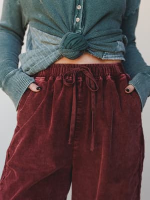 A close-up mid-shot of wide-leg corduroy pants in washed burgundy styled with a front-knotted teal ribbed henley, hands tucked into pockets — the kind of outfit that feels as good as it looks on a slow Saturday.