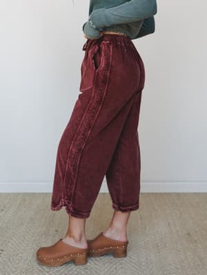 A side-profile shot of wide-leg cropped corduroy pants in a washed burgundy, styled with a cropped teal long-sleeve and brown studded clogs — the kind of effortless fall outfit that looks like you tried just enough.