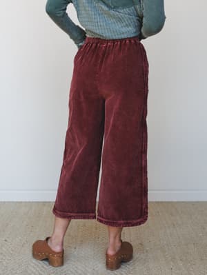 Shot from the back, a model wears wide-leg cropped corduroy pants in a washed burgundy paired with a sage ribbed long-sleeve top and wood-platform clogs — the kind of easy, grounded outfit that works just as hard on a weekend errand run as it does at
