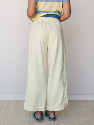 Back view of a model in wide-leg cream pants with a hemmed cuff and elastic waist, styled with a striped crochet top and teal woven sandals against a soft neutral studio wall.