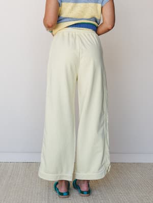 Back view of a model in wide-leg cream pants with a hemmed cuff and elastic waist, styled with a striped crochet top and teal woven sandals against a soft neutral studio wall.