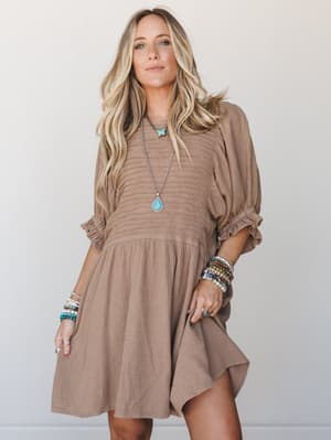A breezy almond-toned mini dress in crinkle gauze with smocked bodice and puff sleeves, styled with stacked turquoise jewelry for that effortless boho-meets-weekend energy.