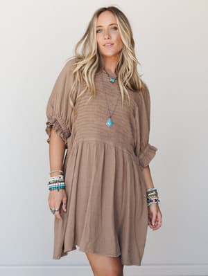 A flowy almond-toned gauze mini dress with smocked bodice and puff sleeves, styled with stacked turquoise and beaded bracelets and a layered pendant necklace — the kind of dress that looks equally at home on a patio or a road trip.