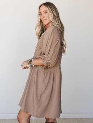 A warm, easygoing mini dress in almond gauze with smocked sleeves and a tiered skirt — styled with a stack of colorful bracelets that feels less 'outfit planned' and more 'this is just how she lives.'