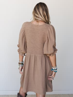 Shot from behind, a model wears a warm almond mini dress with smocked bodice and puff sleeves, styled with stacked boho bracelets and brown boots — effortless and a little romantic.