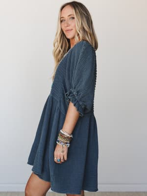 A washed navy gauze mini dress with a relaxed babydoll silhouette and ruched puff sleeves, styled with a stack of colorful beaded bracelets and rings — the kind of easy, expressive outfit that works for both a coffee run and a last-minute dinner plan