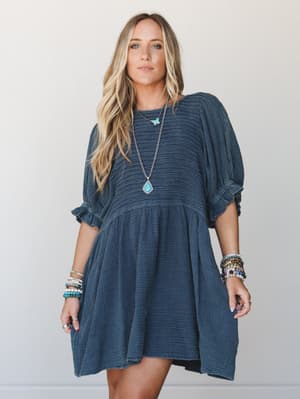 A flowy navy gauze mini dress with a babydoll silhouette, styled with stacked boho bracelets and a turquoise pendant necklace — the kind of effortless outfit that looks like you tried just enough.