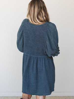 A relaxed mini dress in deep slate blue shot from the back, showing off its smocked bodice, billowy puff sleeves, and gathered skirt — the kind of dress that looks effortless but still feels like a whole outfit.